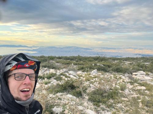 Henry on one of his bike rides, at the top of a mountain with a view of the sky and land behind him 